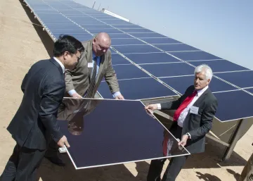 Three men in business suits lifting a solar panel onto the larger collection of solar panels together.